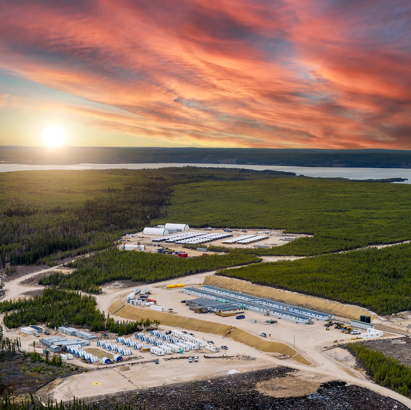 Aerial view of a remote construction site with buildings surrounded by green forest under a colorful sunset.