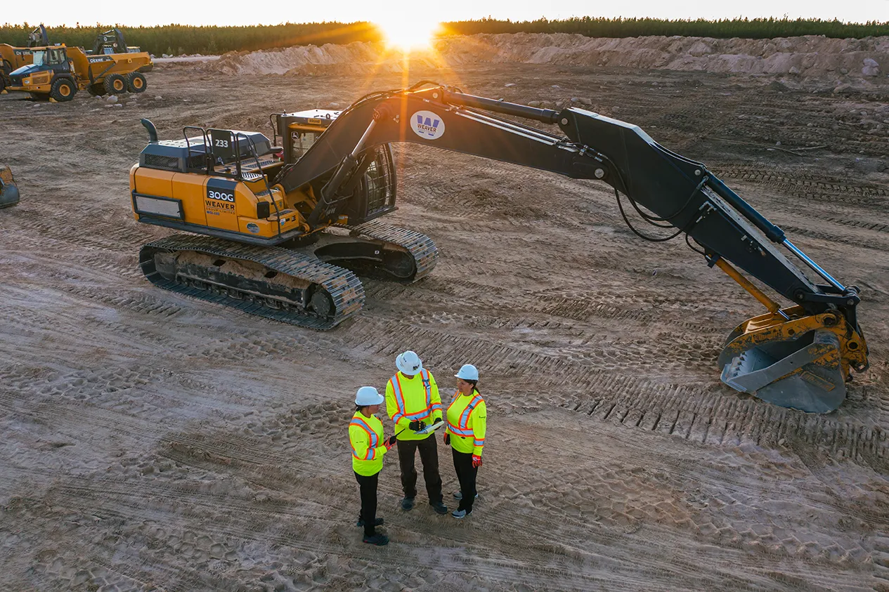 Three construction workers in safety gear standing on a dirt site next to a large excavator at sunset.