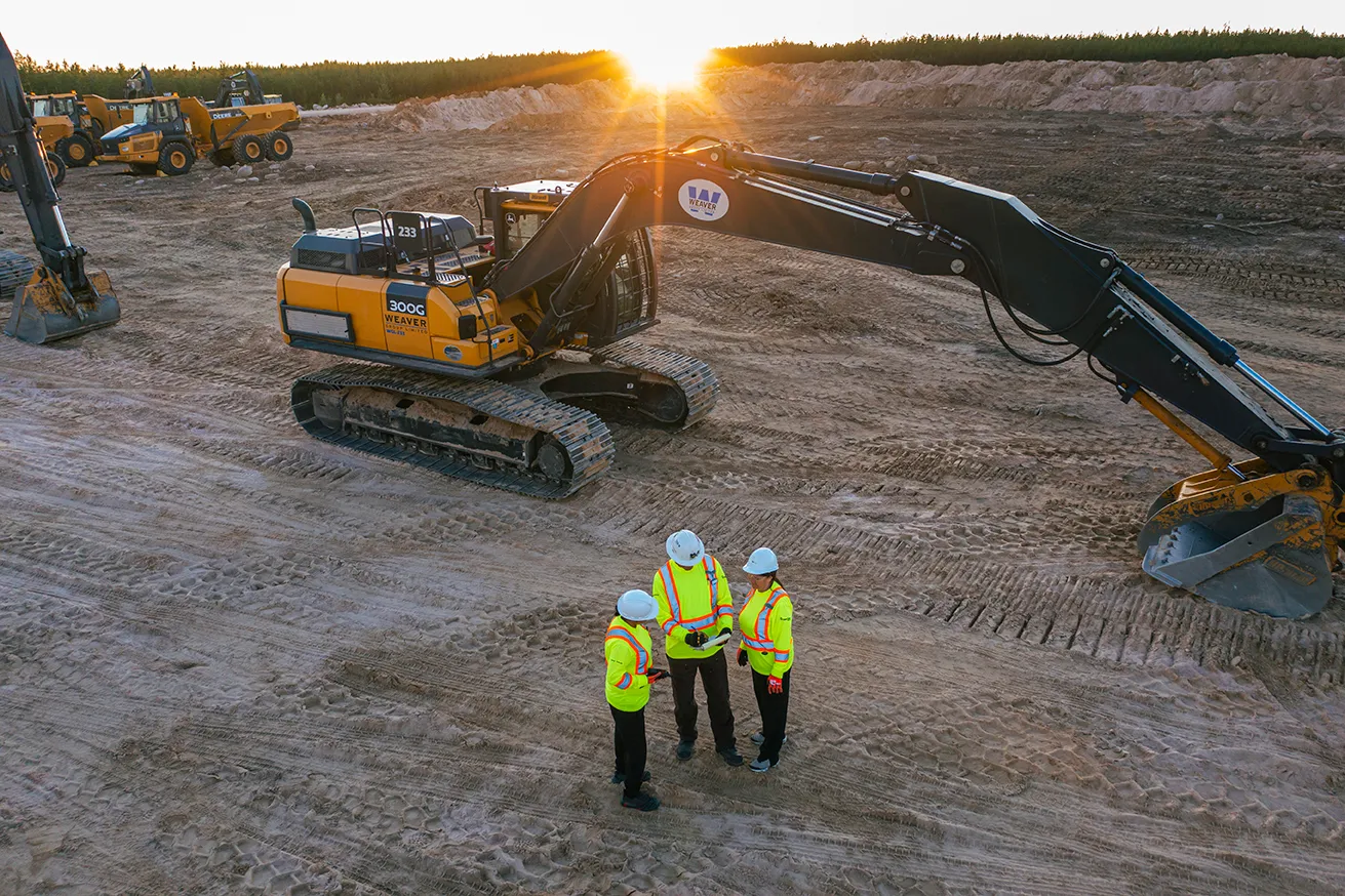 Three construction workers in neon jackets and helmets standing on dirt near large excavators at sunset.