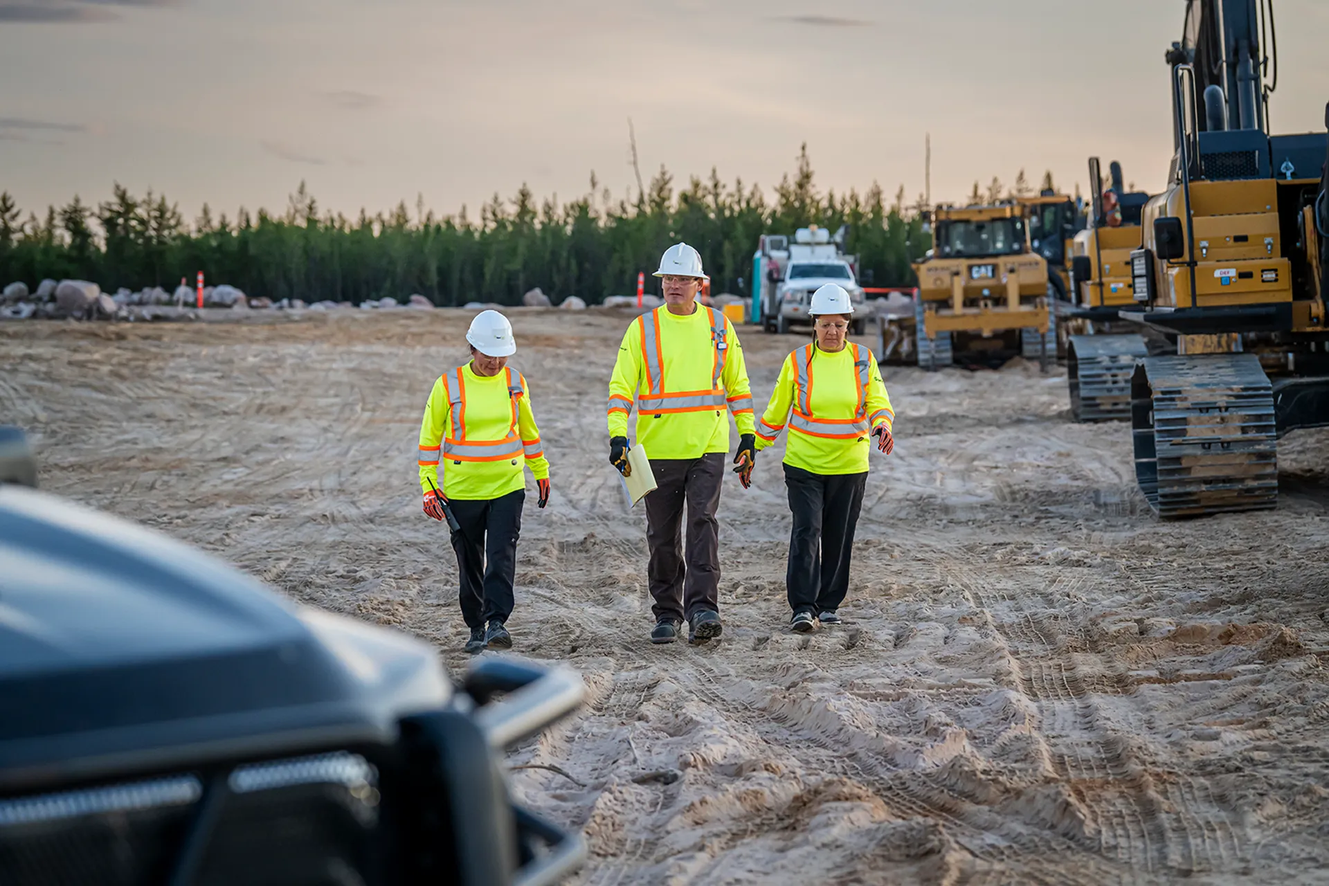 Three construction workers in yellow safety gear walking on a dirt site with machinery in the background.