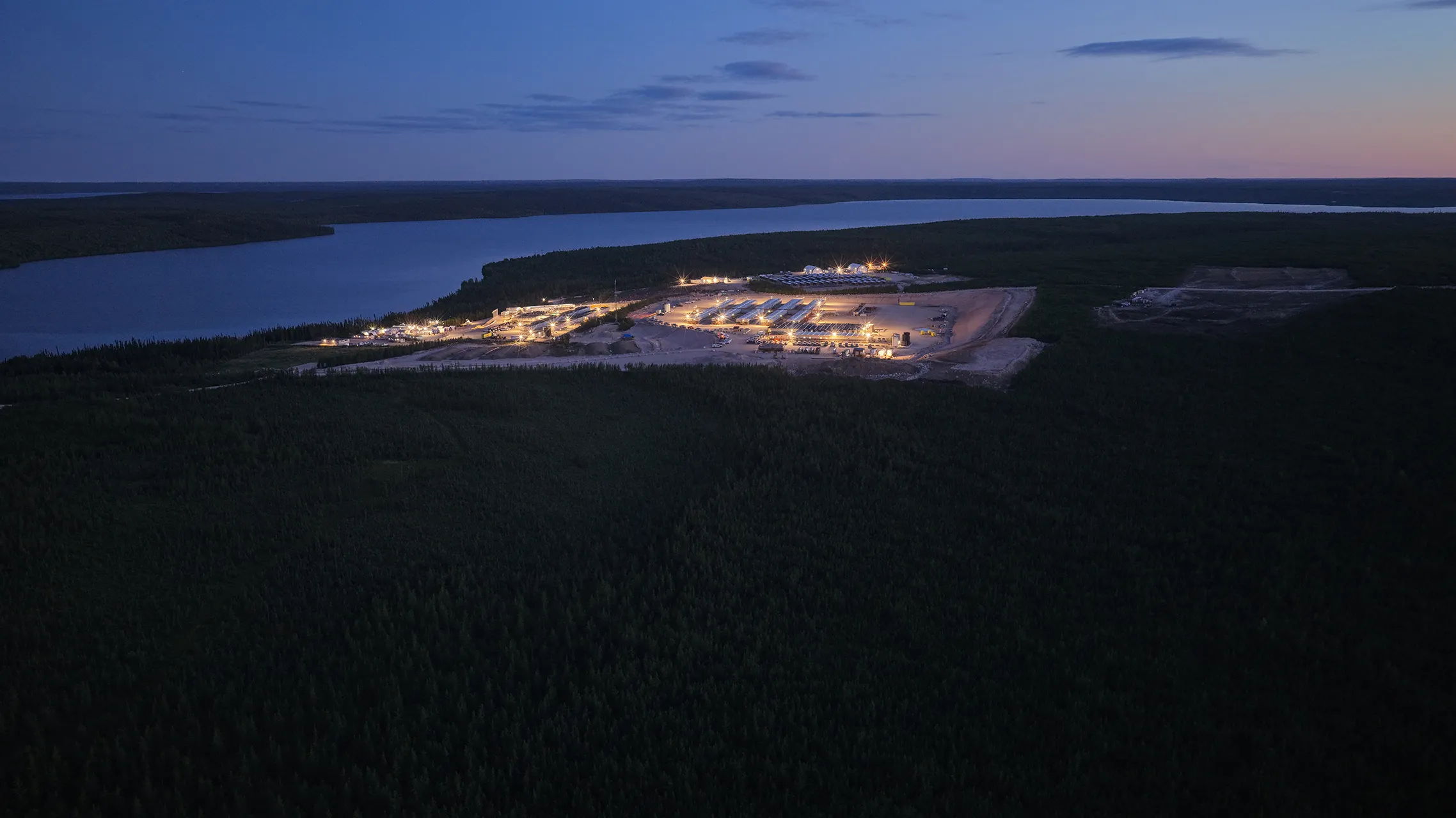 Aerial nighttime view of an illuminated industrial site near a large river surrounded by dense forest.