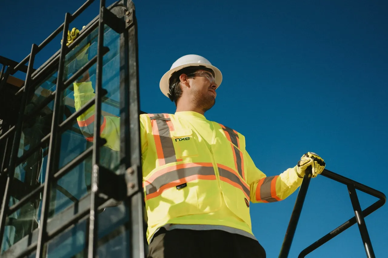 Construction worker in a hard hat and safety vest stands on machinery stairs against a clear blue sky.