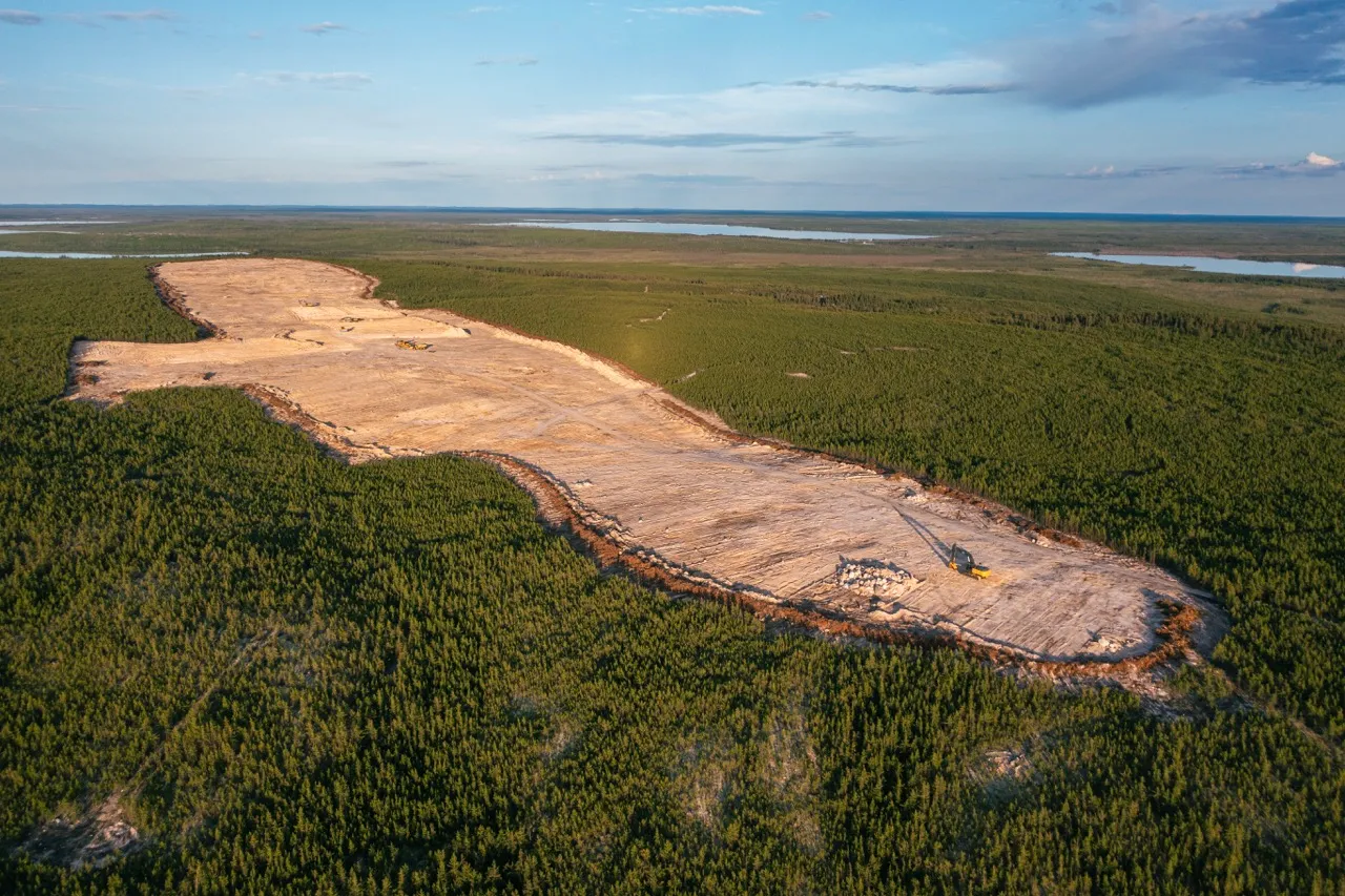 Large cleared land area with construction vehicles surrounded by dense green forest and lakes in the background.