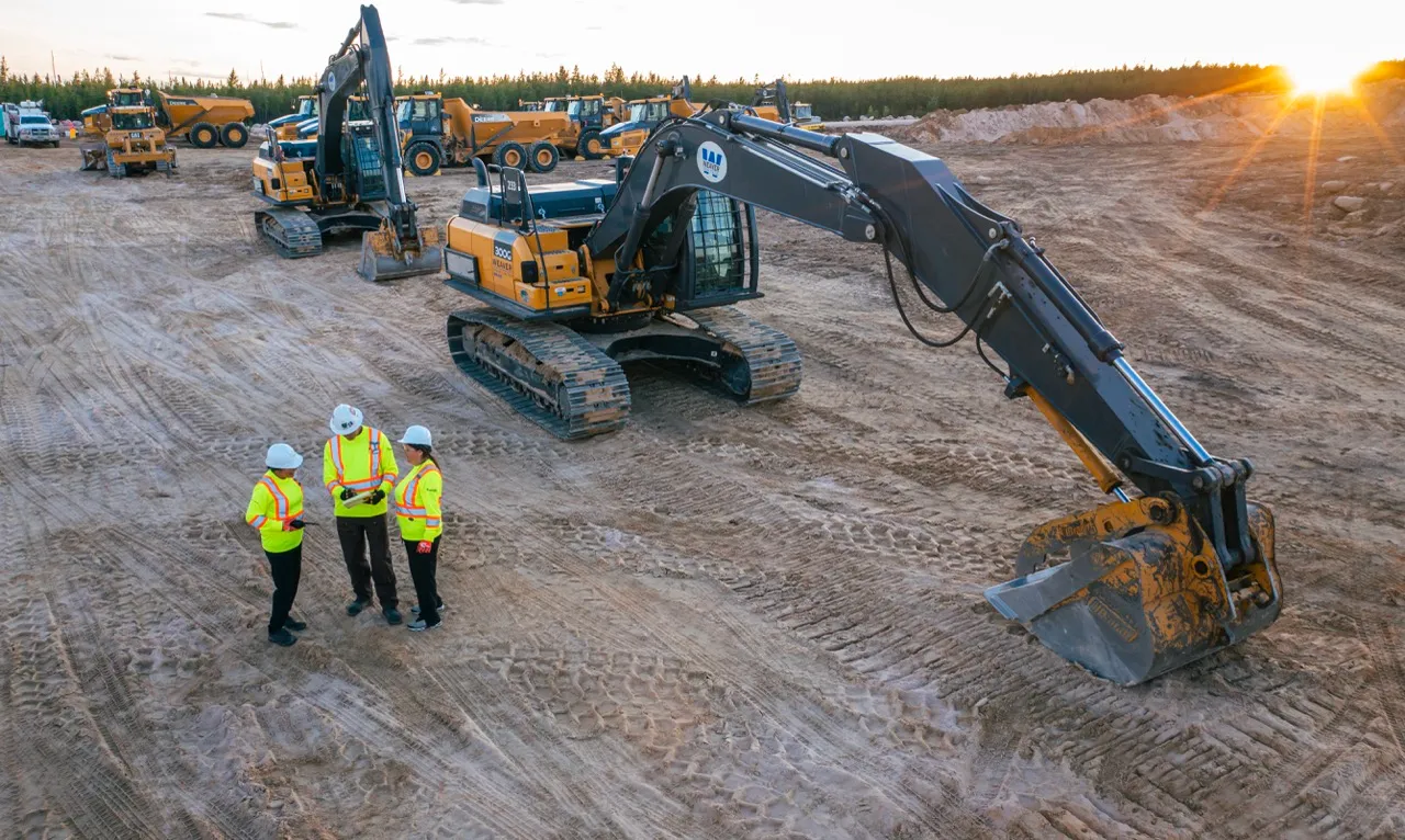 Three construction workers in safety gear standing by excavators and trucks on a dirt site at sunset.