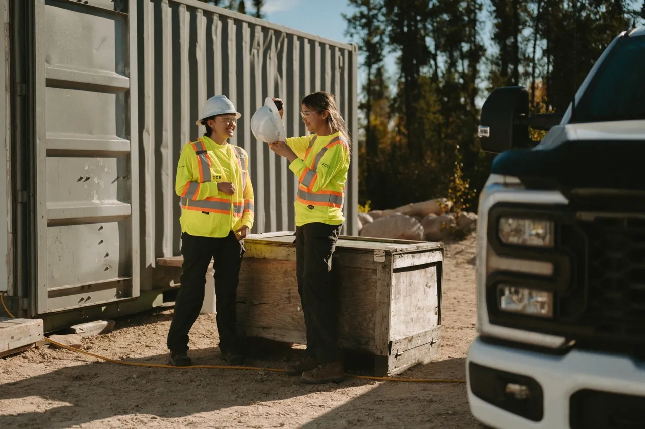 Two female construction workers in yellow safety shirts and helmets standing by equipment outdoors.