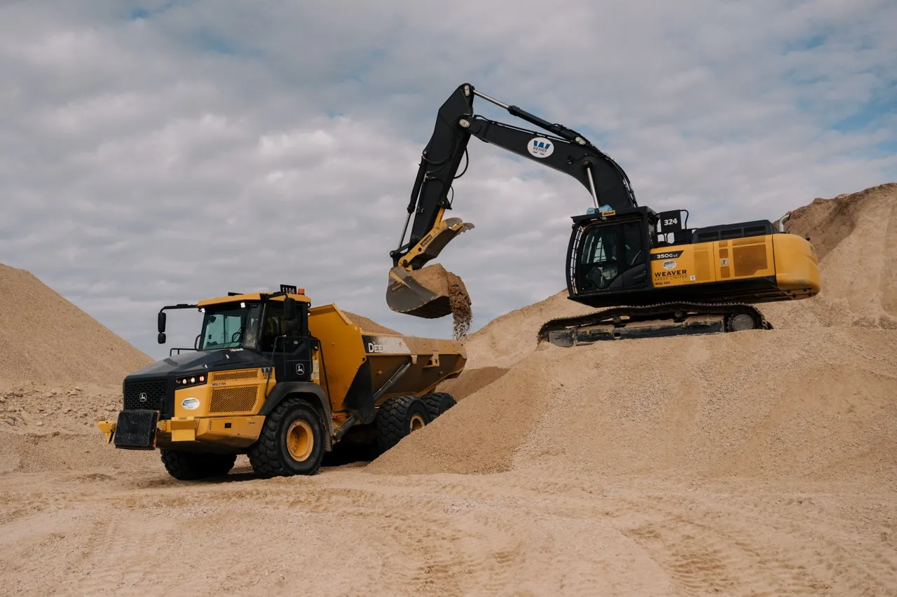 Yellow excavator loading sand into a yellow dump truck in a sandy construction site.