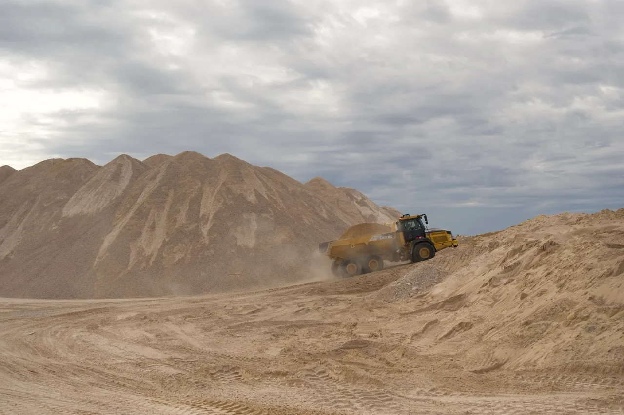 Yellow dump truck carrying sand uphill at a sandy construction or mining site under cloudy sky.