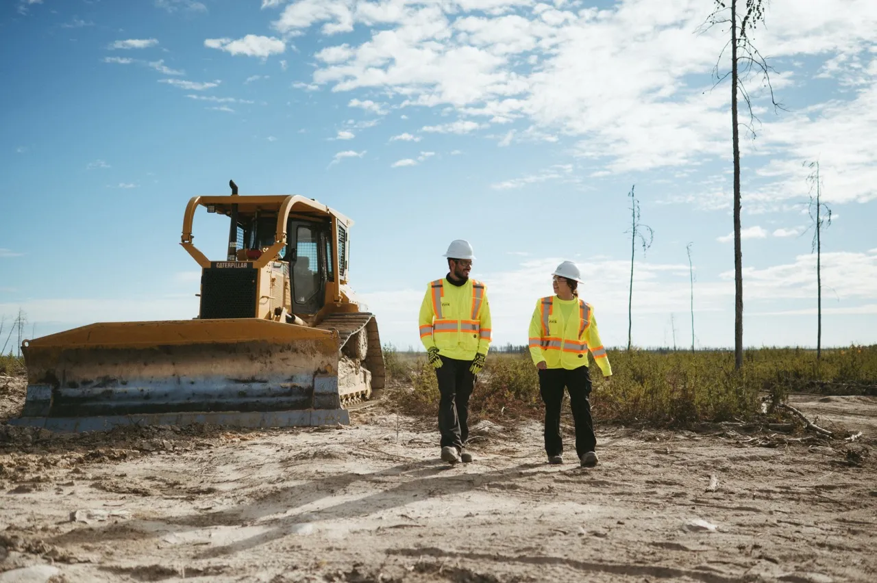 Two workers in safety gear walking past a large yellow bulldozer on a clear day.