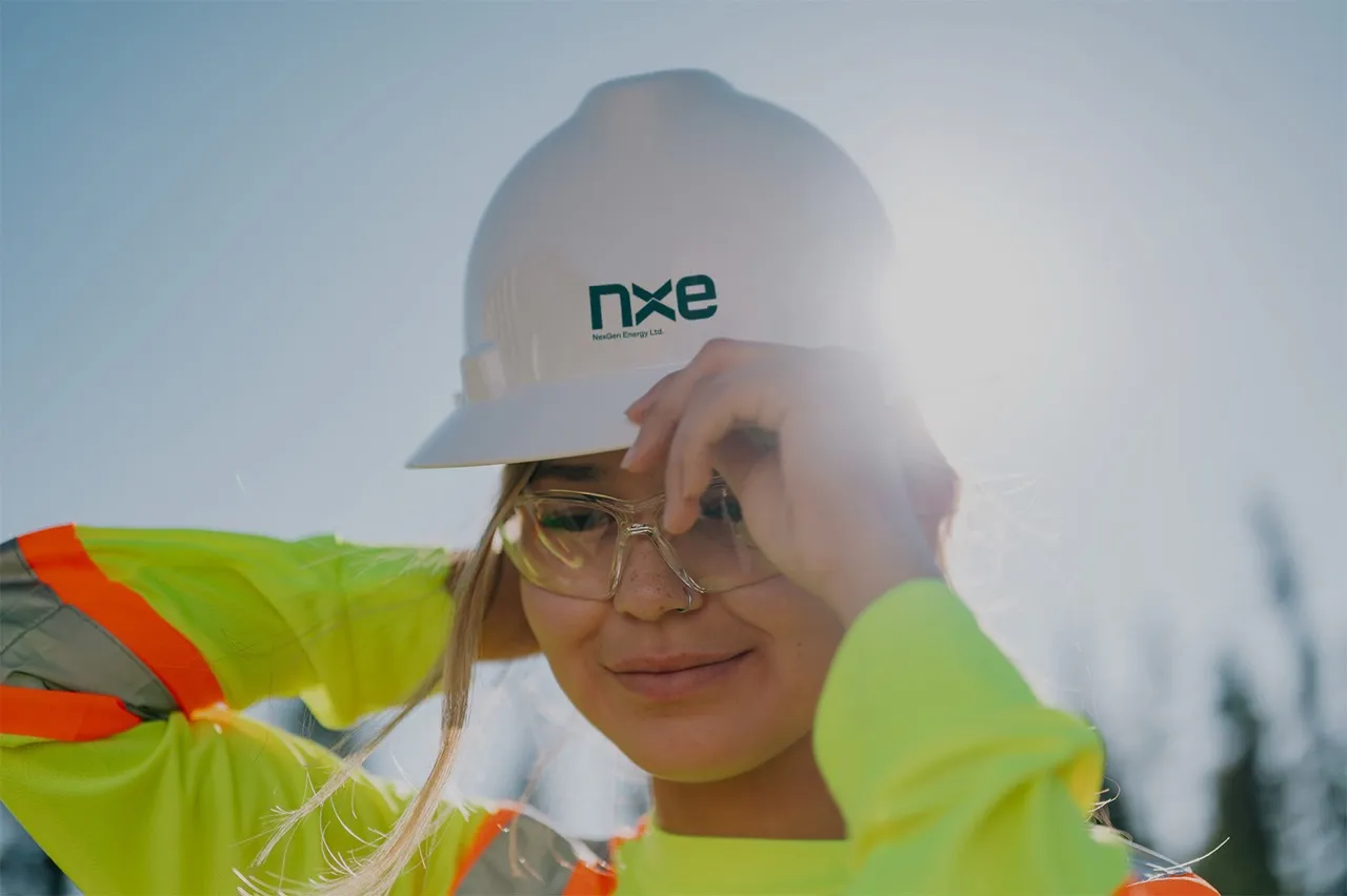 Woman in bright yellow safety gear adjusting white hard hat with Nxe logo outdoors in daylight.