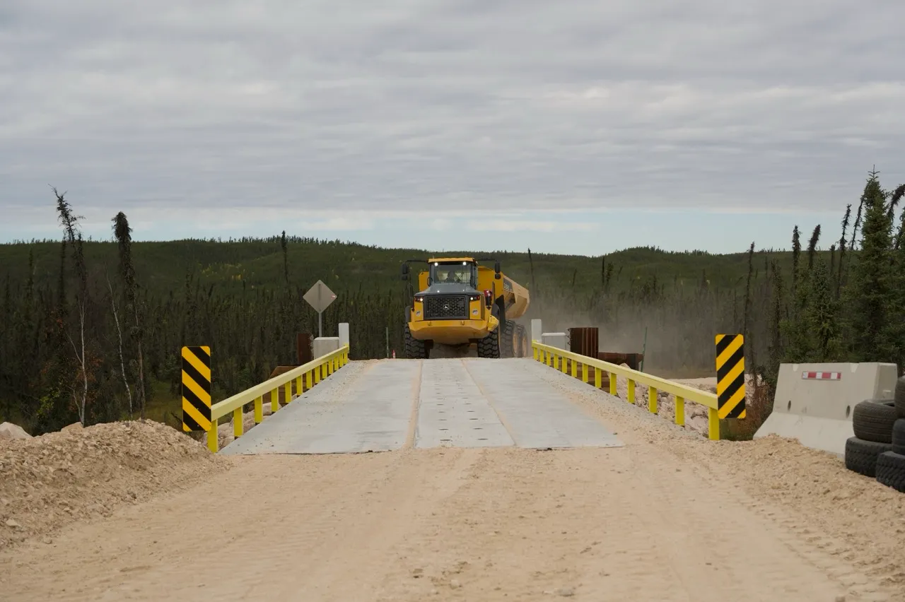 Yellow construction truck driving on a dirt road crossing a small bridge with yellow railings.