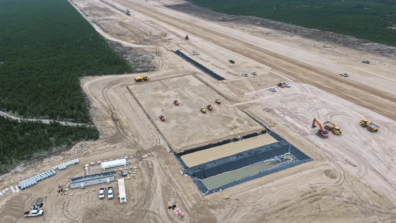 Aerial view of a large construction site with heavy machinery preparing a wide dirt airstrip surrounded by forest.