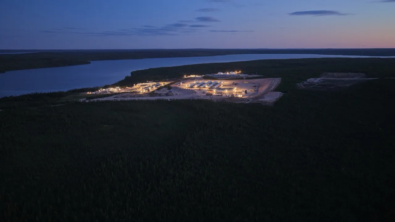 A lit industrial site at dusk surrounded by dense forest near a wide river under a darkening sky.