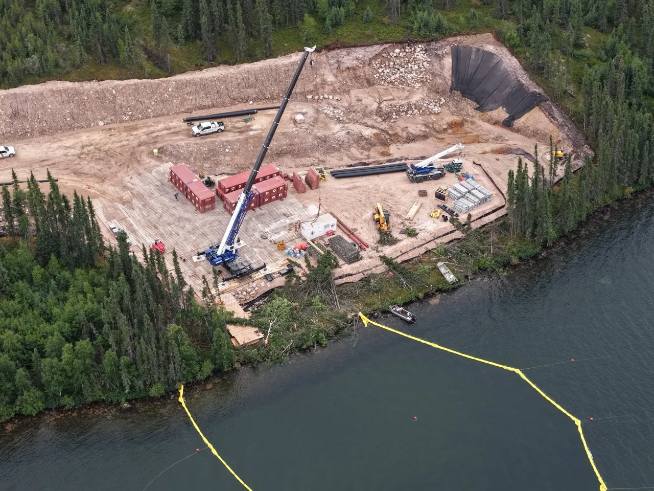 Crane lifting a concrete structure on a clear pad by a lakeshore surrounded by forest and equipment.