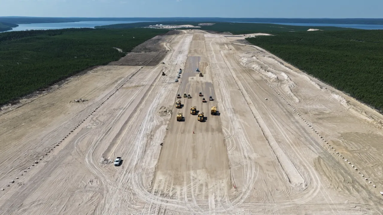 Aerial view of construction vehicles compacting and preparing a long dirt airstrip surrounded by forest.