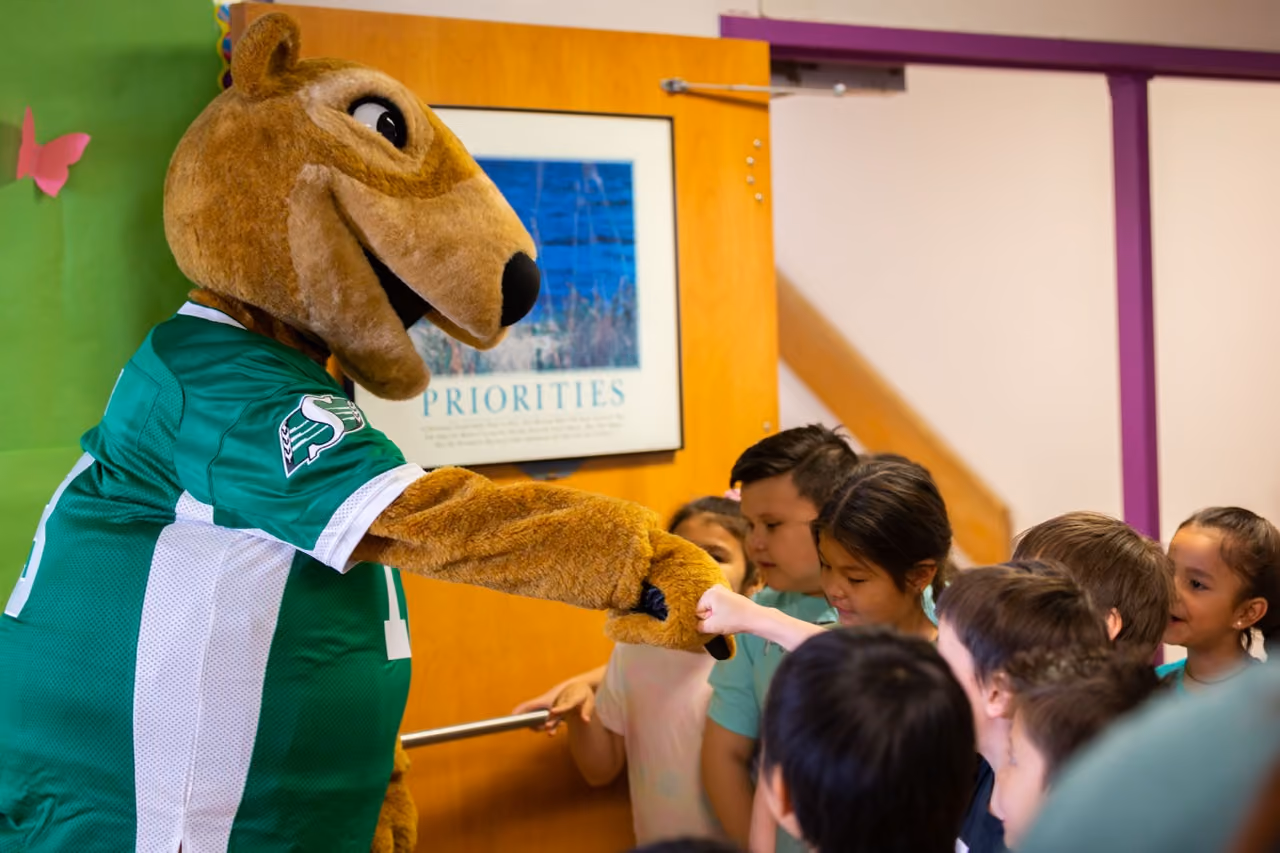 Sports mascot wearing green jersey fist bumps a group of children inside a room.
