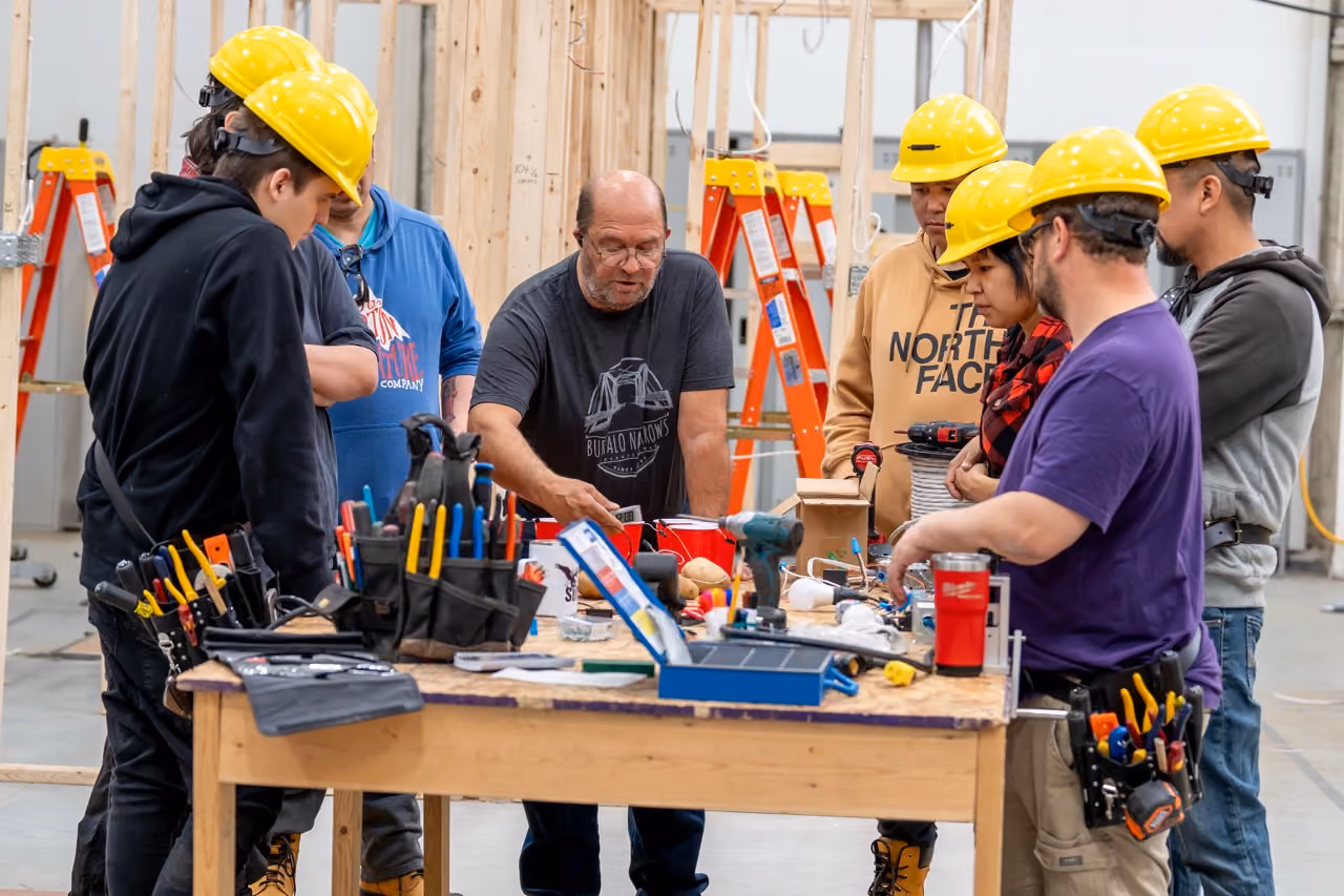 Group wearing yellow hard hats gathered around a workbench with tools in a construction setting.