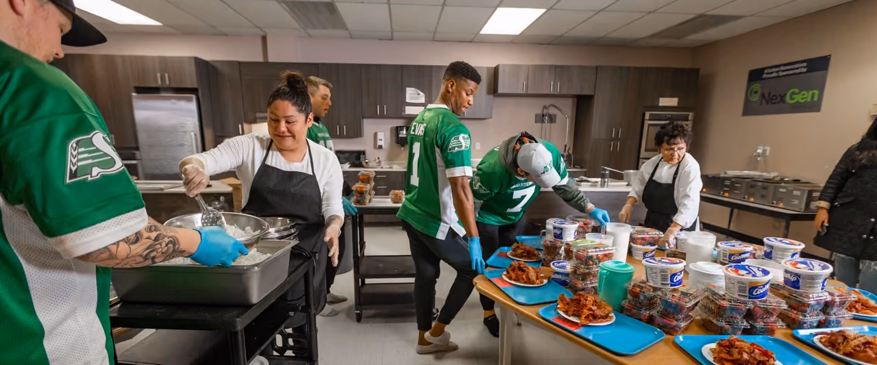 Volunteers wearing green jerseys and aprons prepare and serve food in a kitchen with trays of bacon and containers.