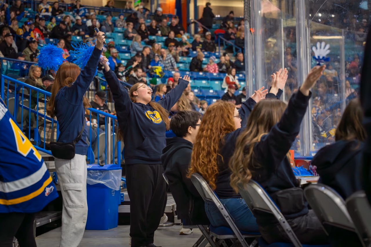 Cheering young fans with blue pom-poms and raised arms at an indoor sports event with seated crowd.