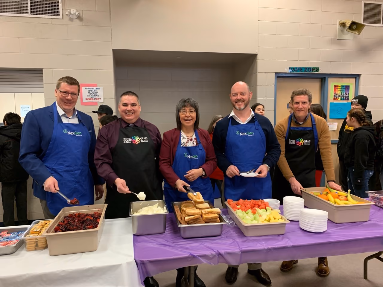 Five people wearing aprons serving breakfast foods like fruit, toast, and yogurt in a school cafeteria.