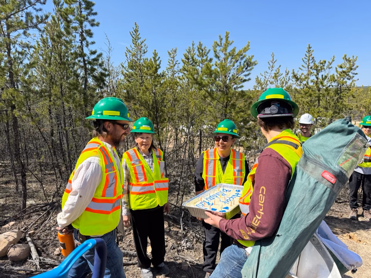 Group of workers wearing green helmets and safety vests outdoors with one holding a cake tray.