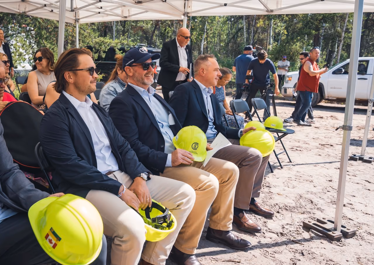 Men in business attire holding yellow hard hats seated under a tent at an outdoor event.