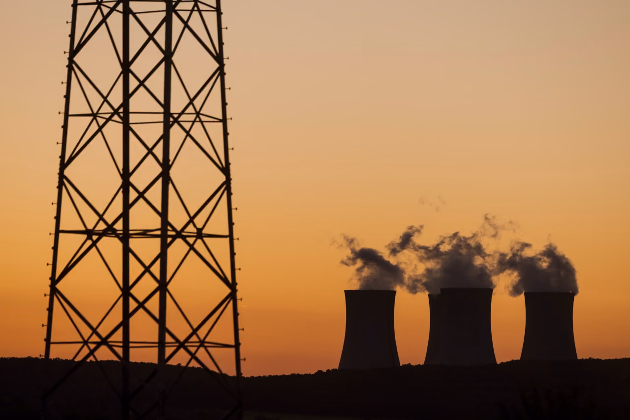 Silhouette of three cooling towers emitting steam at sunset with a tall metal structure in the foreground.