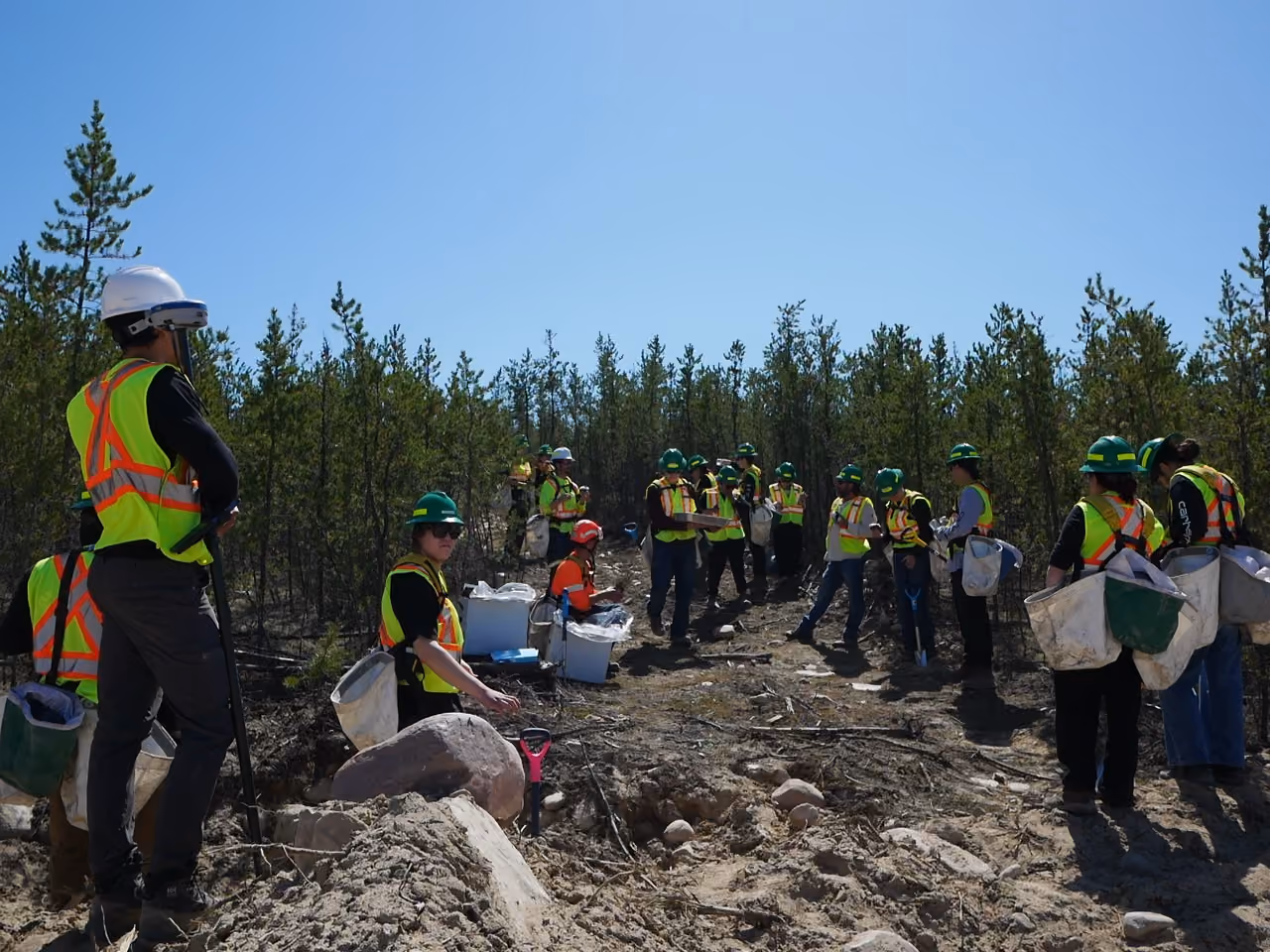 Group of workers in safety vests and helmets standing and sitting outdoors on a dirt path near trees.