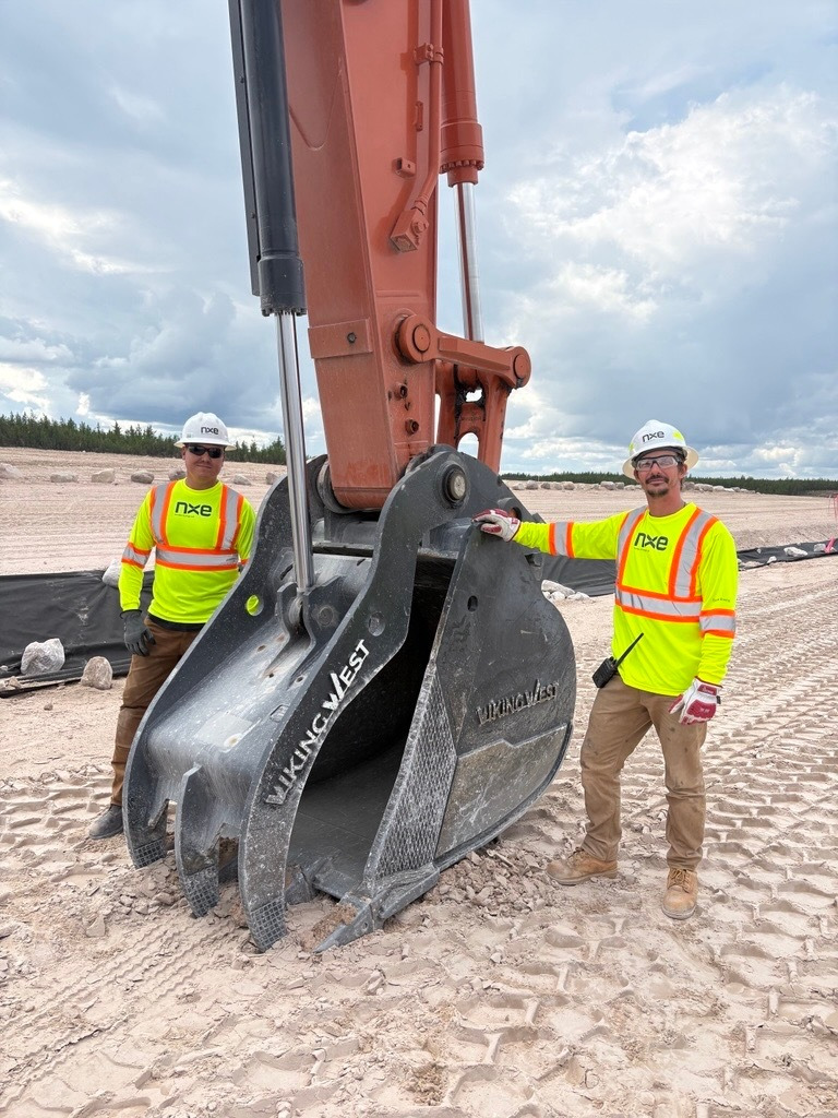 Two construction workers in neon safety shirts and helmets stand by a large excavator bucket on a dirt site.