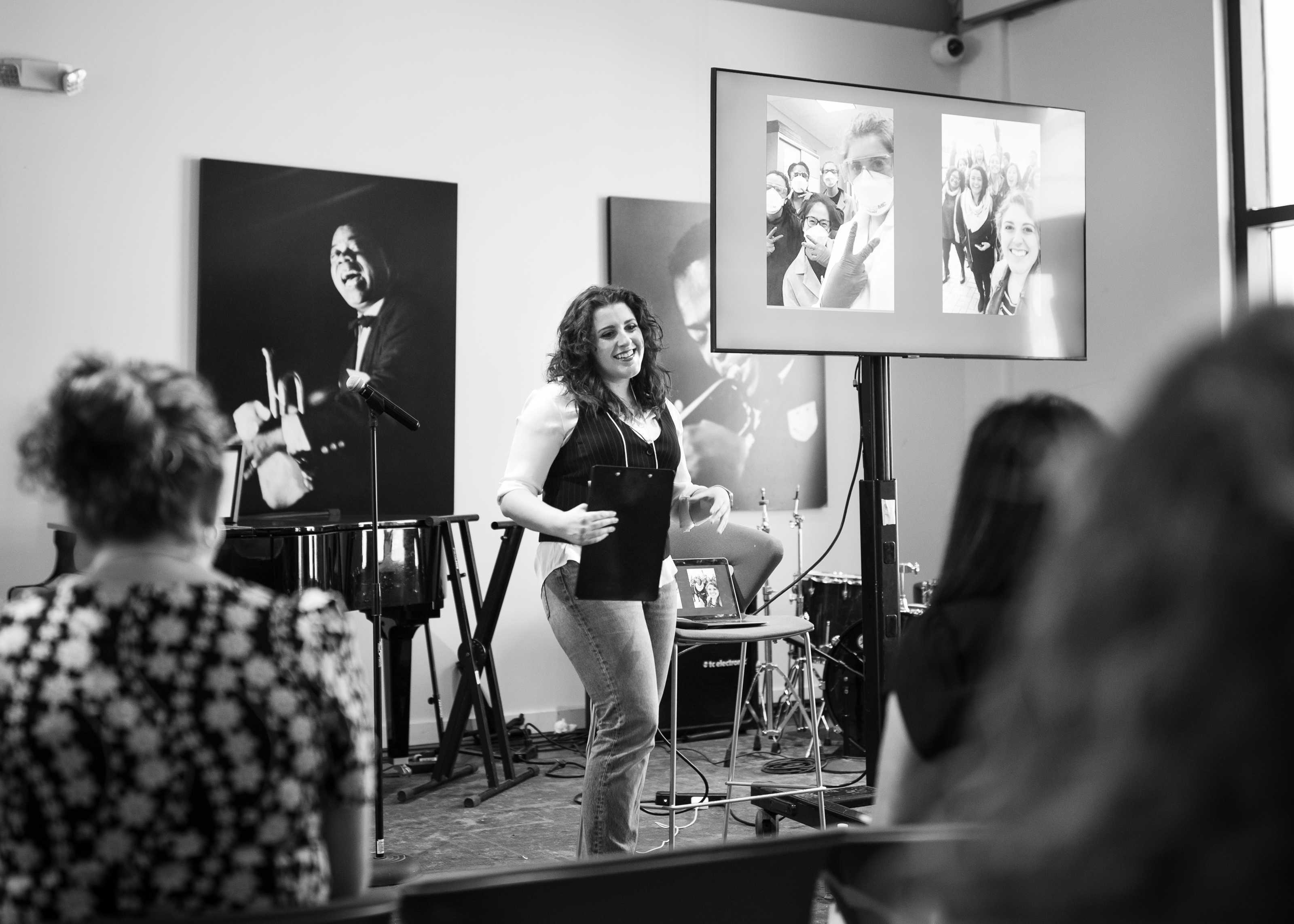 Photo of a woman with curly hair holding a clipboard standing in front of an audience. A tv screen is to her right displaying photos.