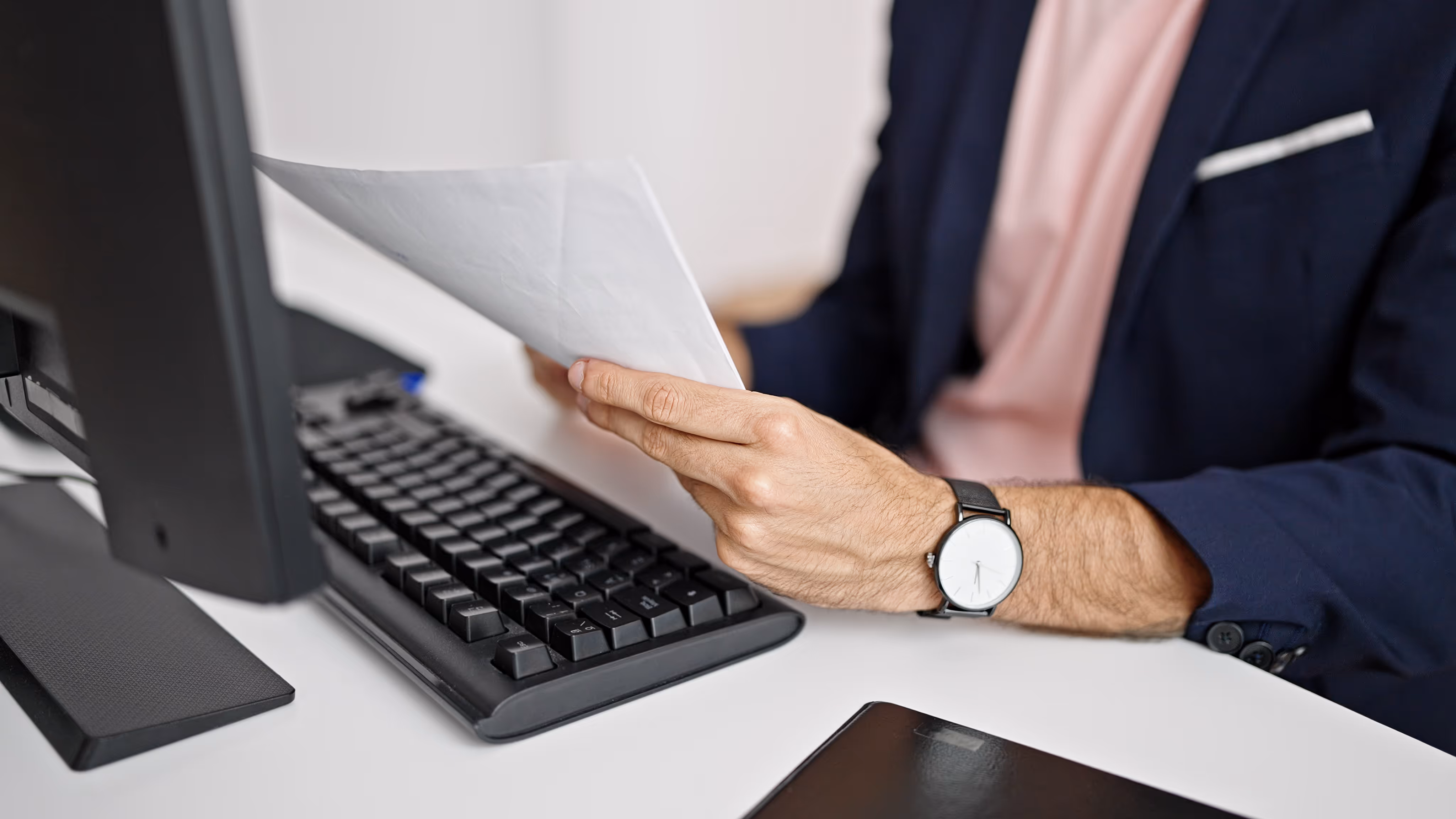 Person in business attire holding and reviewing papers at a desk with a keyboard and monitor.