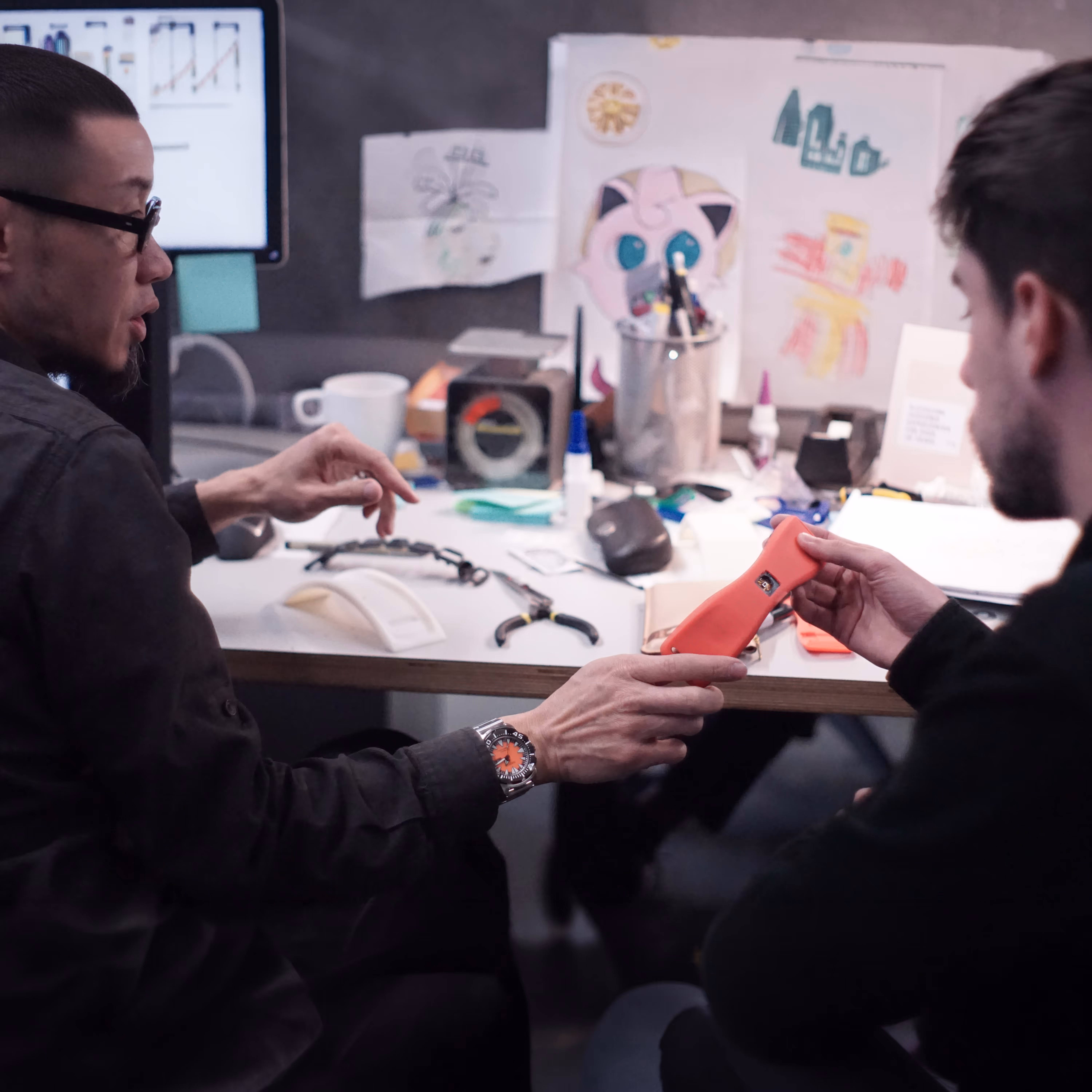 Two men seated at a desk discussing and holding a red prototype device with various tools and sketches in the background for product design
