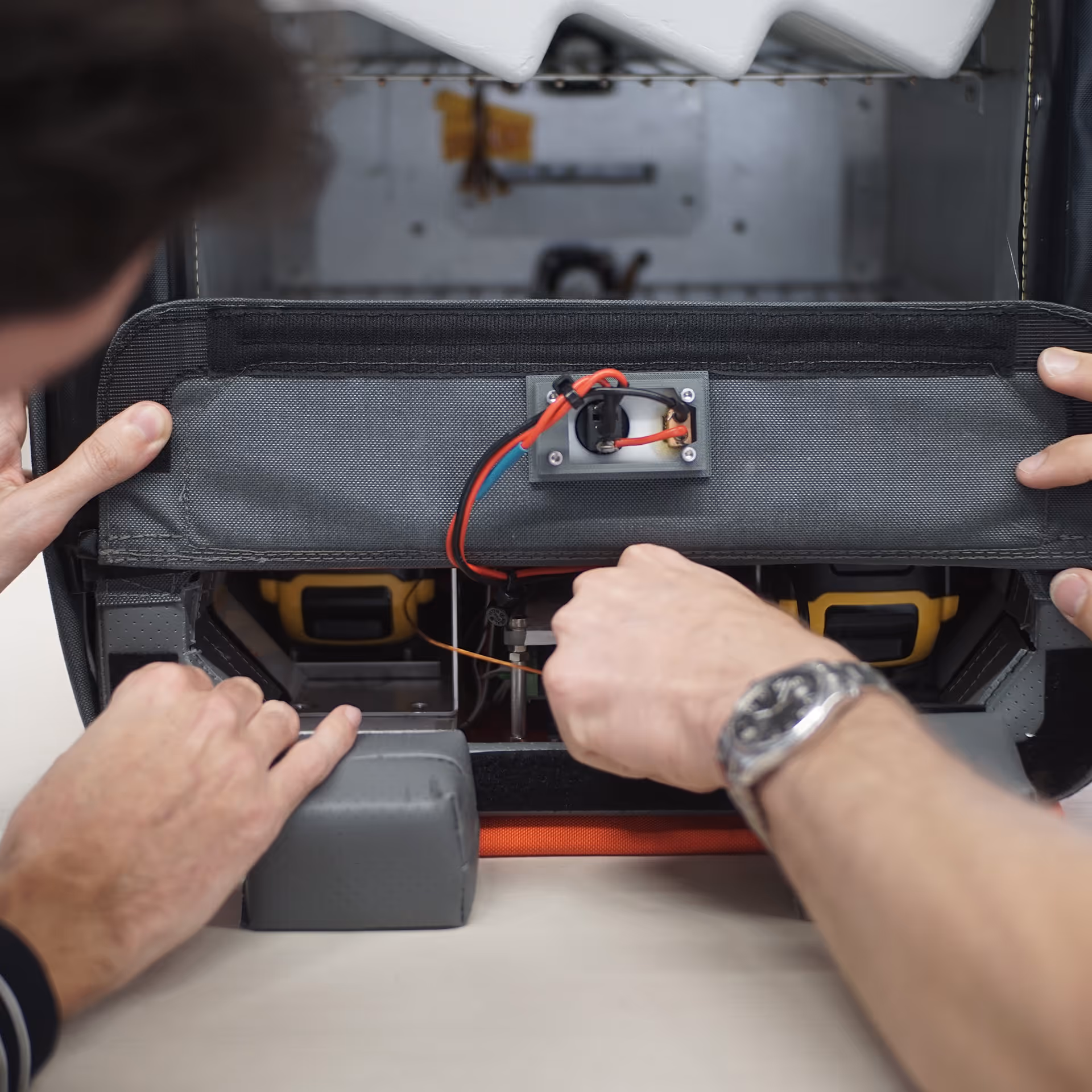 Two hands adjusting wiring inside a gray fabric panel with red and blue wires visible.