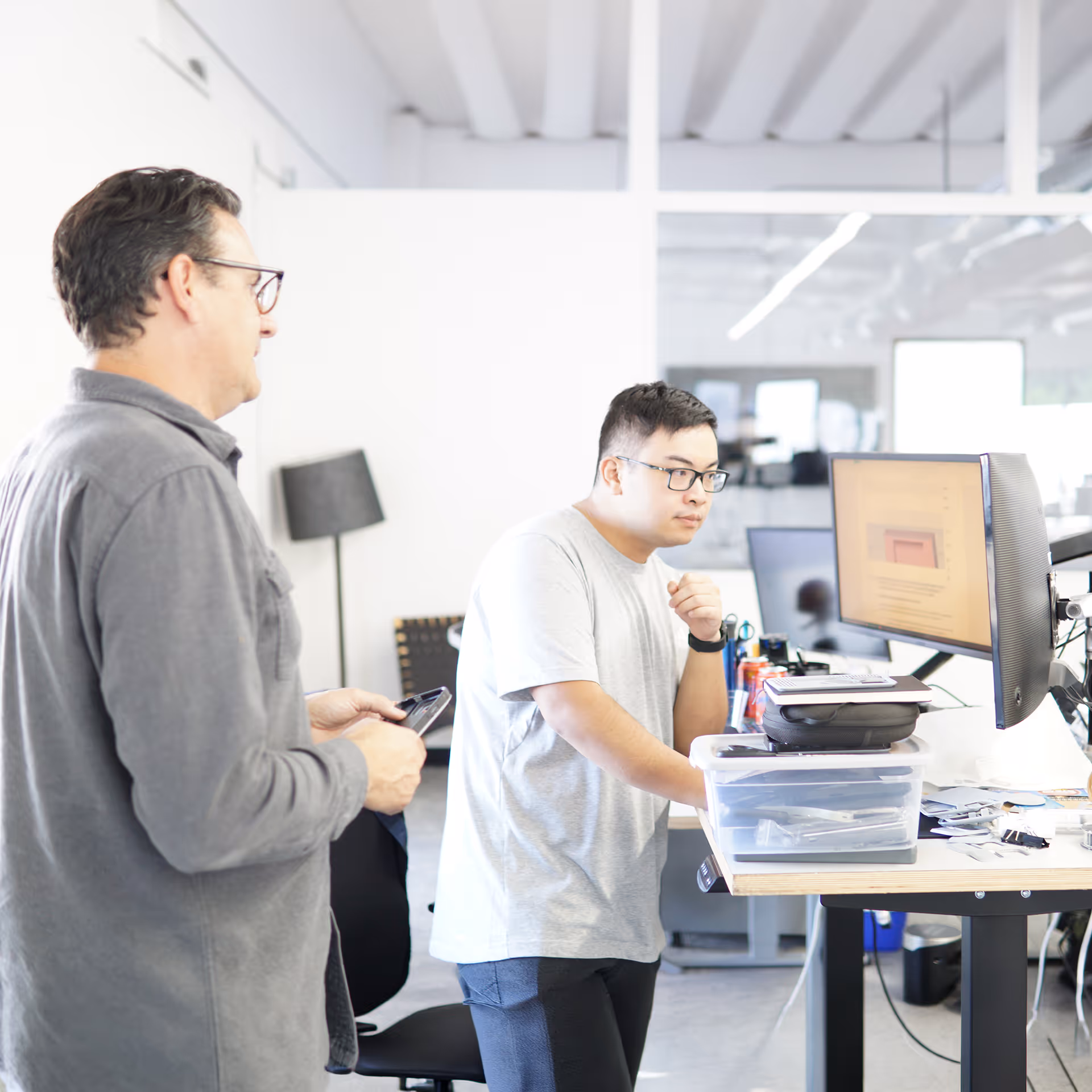 Two men in glasses working at a standing desk with a computer monitor in a bright office.
