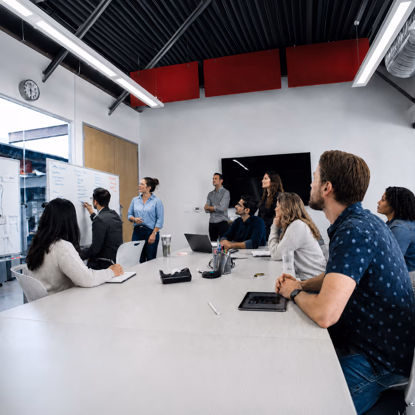 A group of eight people in a meeting room, one writing on a whiteboard while others watch and listen.