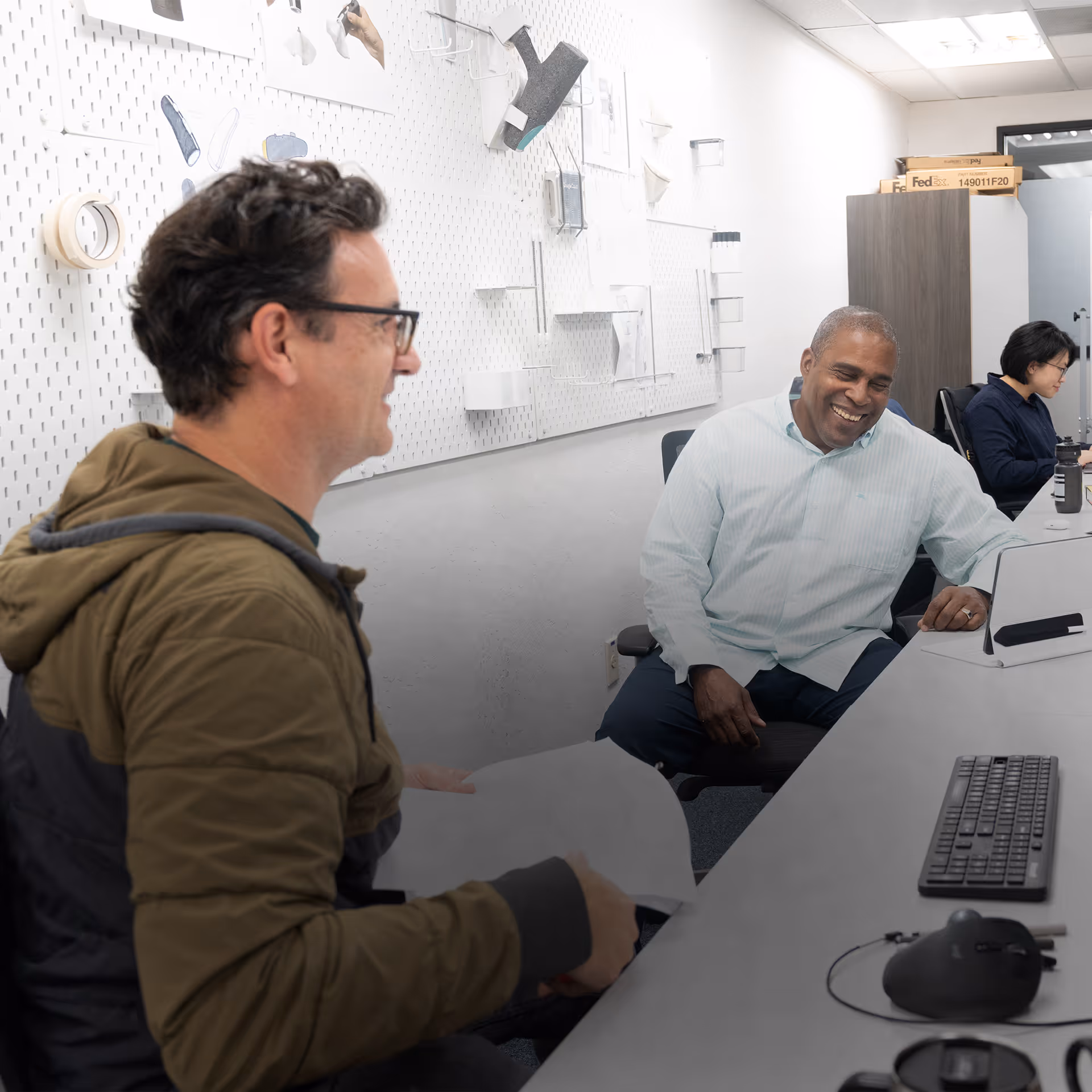 Two men smiling and talking at a desk in an office while a woman works on a computer nearby.