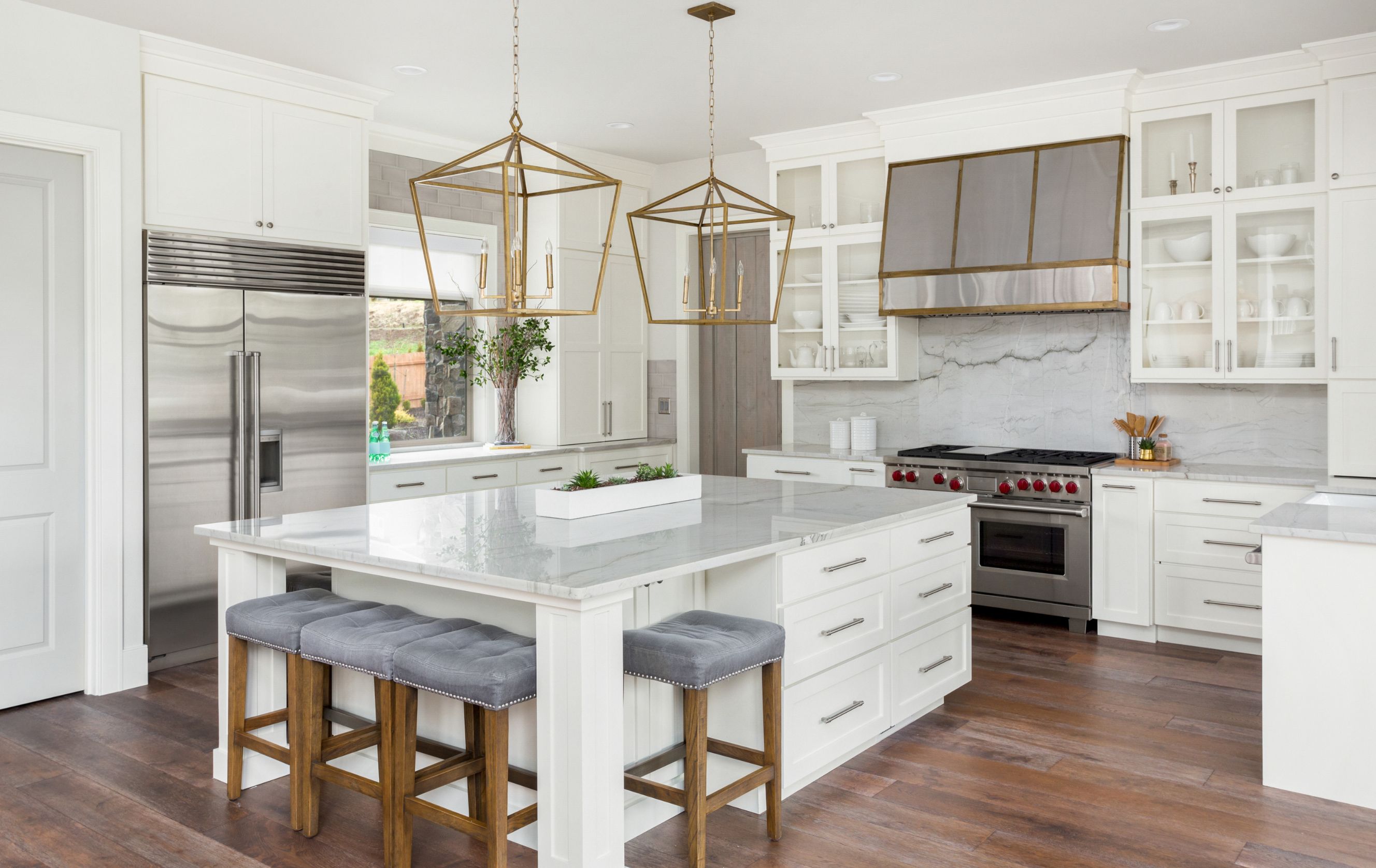 Spacious modern kitchen with white cabinetry, marble island, wooden stools with gray cushions, stainless steel appliances, and gold geometric pendant lights.