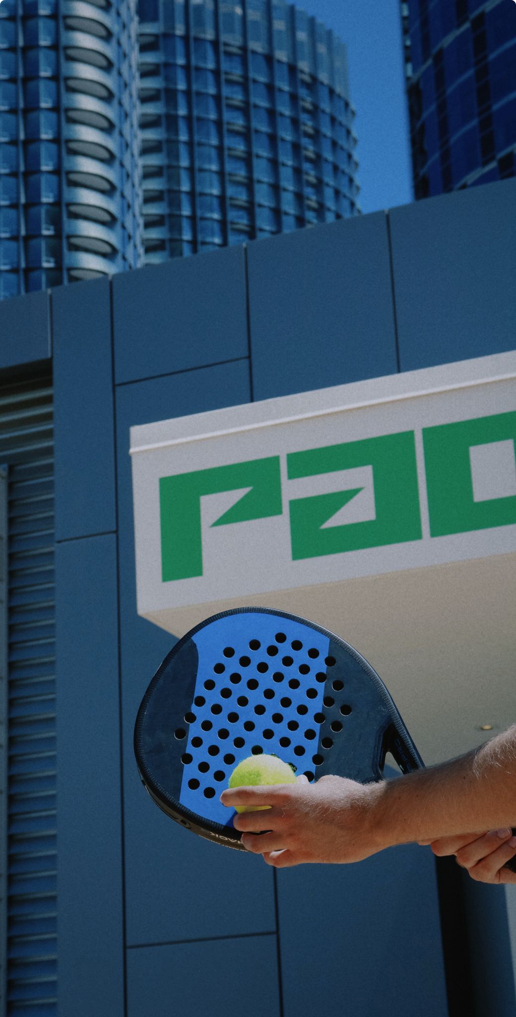 Close-up of a hand holding a padel racket and yellow tennis ball with modern buildings in the background.