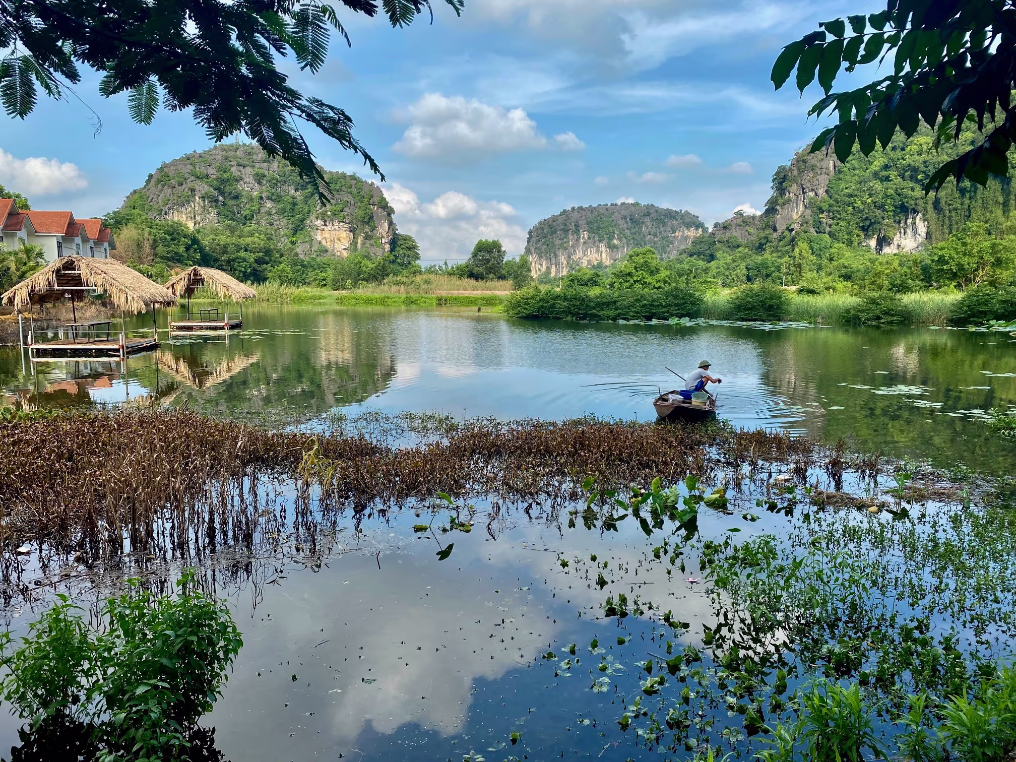 Un lac idyllique entouré d'une végétation luxuriante et de falaises calcaires caractéristiques dans un paysage paisible. Au premier plan, une personne traverse l'eau à bord d'un petit bateau en bois, tandis qu'à l'arrière-plan, des cabanes au toit de chaume se trouvent sur le rivage. Le reflet du ciel bleu et des rochers dans l'eau renforce l'atmosphère paisible.