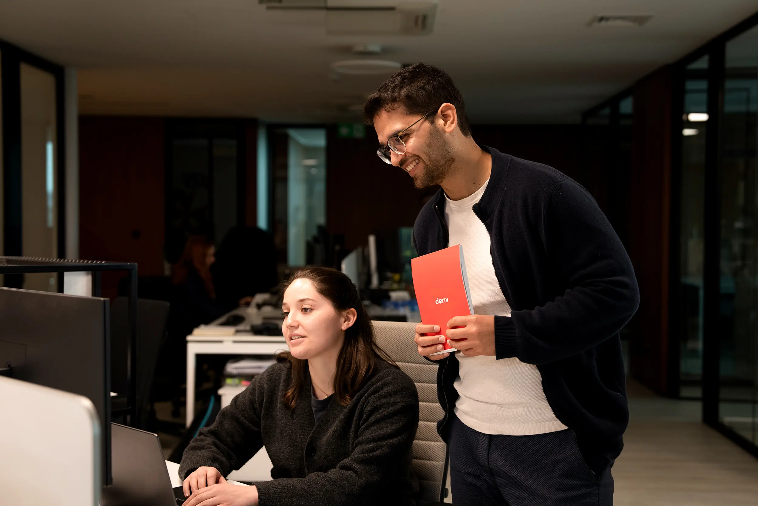 Two teammates collaborating in an office, one holding a red Deriv notebook representing AI innovations in trading.