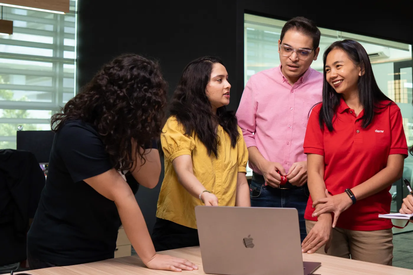 Four Deriv employees collaborating around a laptop, discussing AI innovations to enhance trading performance.