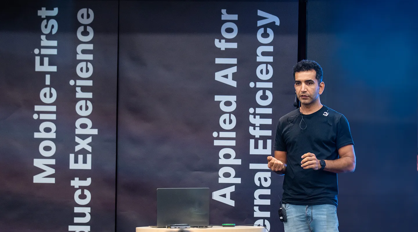 Deriv employee presenting at company event with backdrop displaying core values: AI-first, client-oriented, applied AI for fintech, and operational efficiency