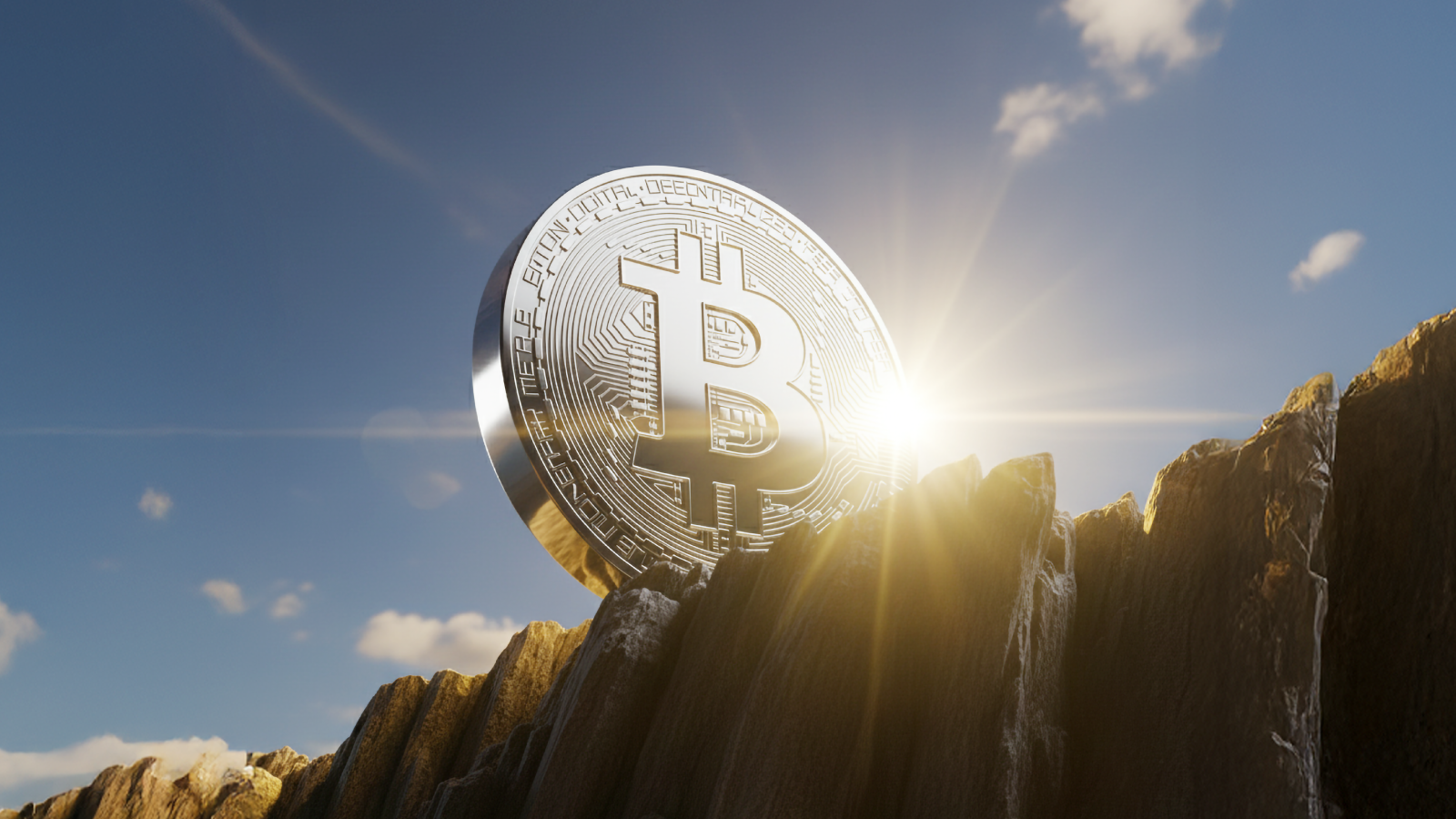 A shiny silver Bitcoin coin standing upright on rocky terrain with sunlight shining behind it, symbolising Bitcoin’s strength and rising value against a clear blue sky.