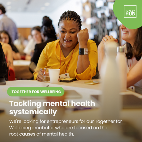 A photograph focusing on one woman sitting at a table in a professional environment, smiling and looking at a document on the table. Around her and in the background are other people are seated in friendly discussion, The Impact Hub logo is placed at the right right hand side of the image, against a light green background. In the bottom half of the image is the text: 'Together for wellbeing. Tackling mental health systemically. We're looking for entrepreneurs for our Together for Wellbeing incubator who are focused on the root causes of mental health.
