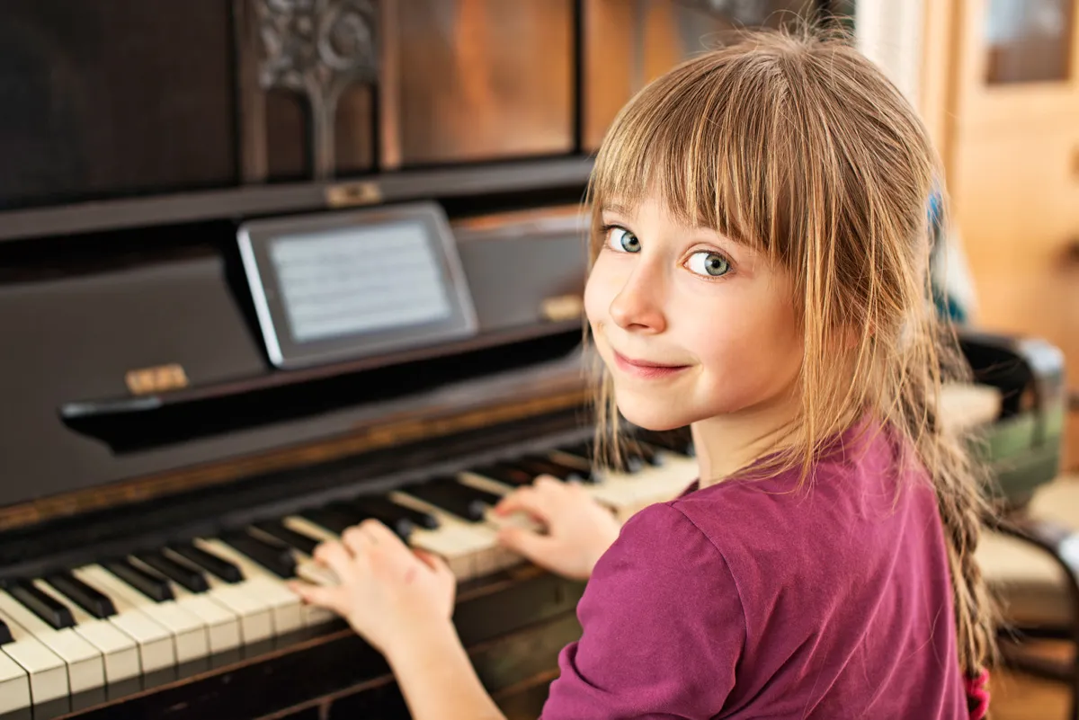 A young girl with light brown hair and bangs, wearing a magenta top, sits at an upright piano with her hands on the keys, looking back at the camera with a smile. A tablet displaying sheet music sits on the piano's music stand.