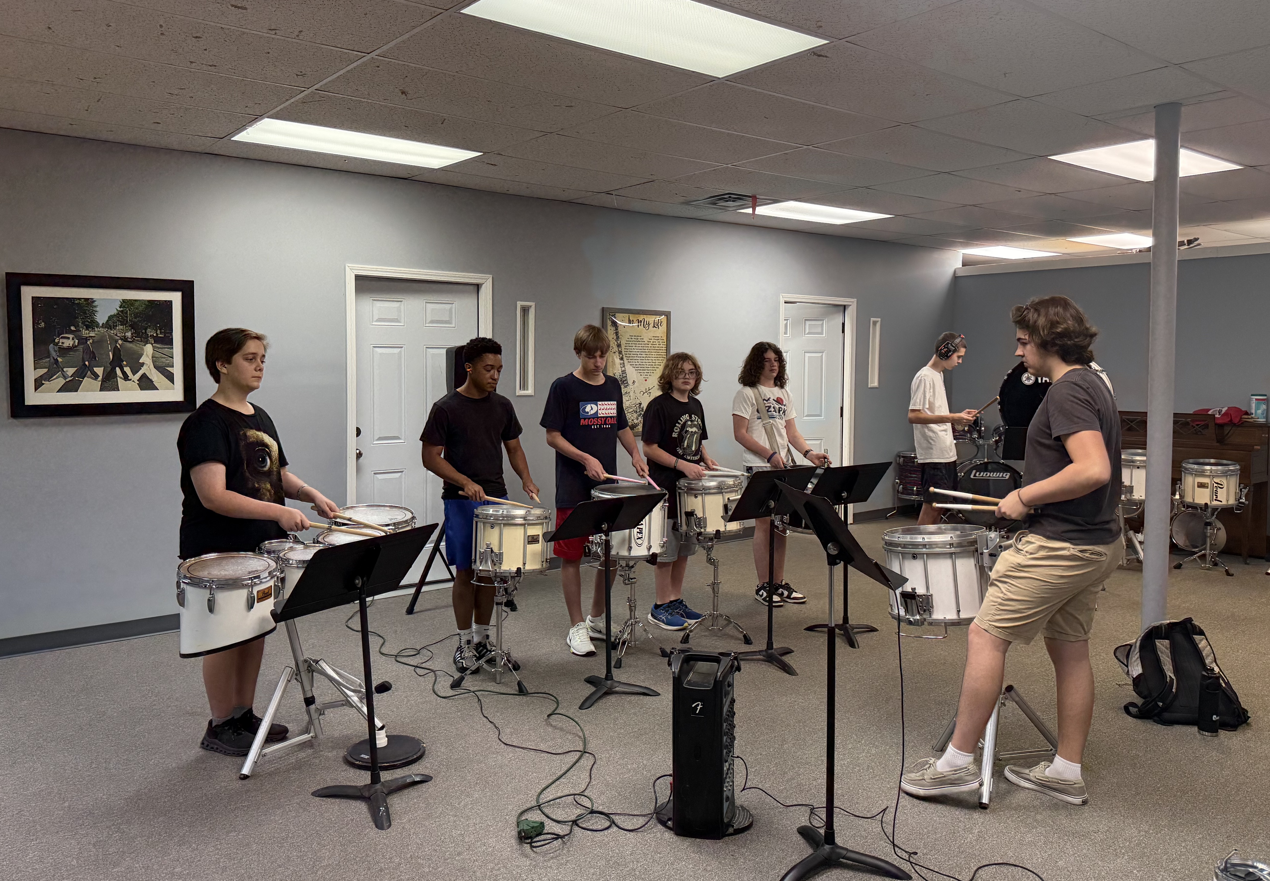 A row of teenage students play matching snare drums together with sheet music stands in a rehearsal room, with an Abbey Road poster on the wall.