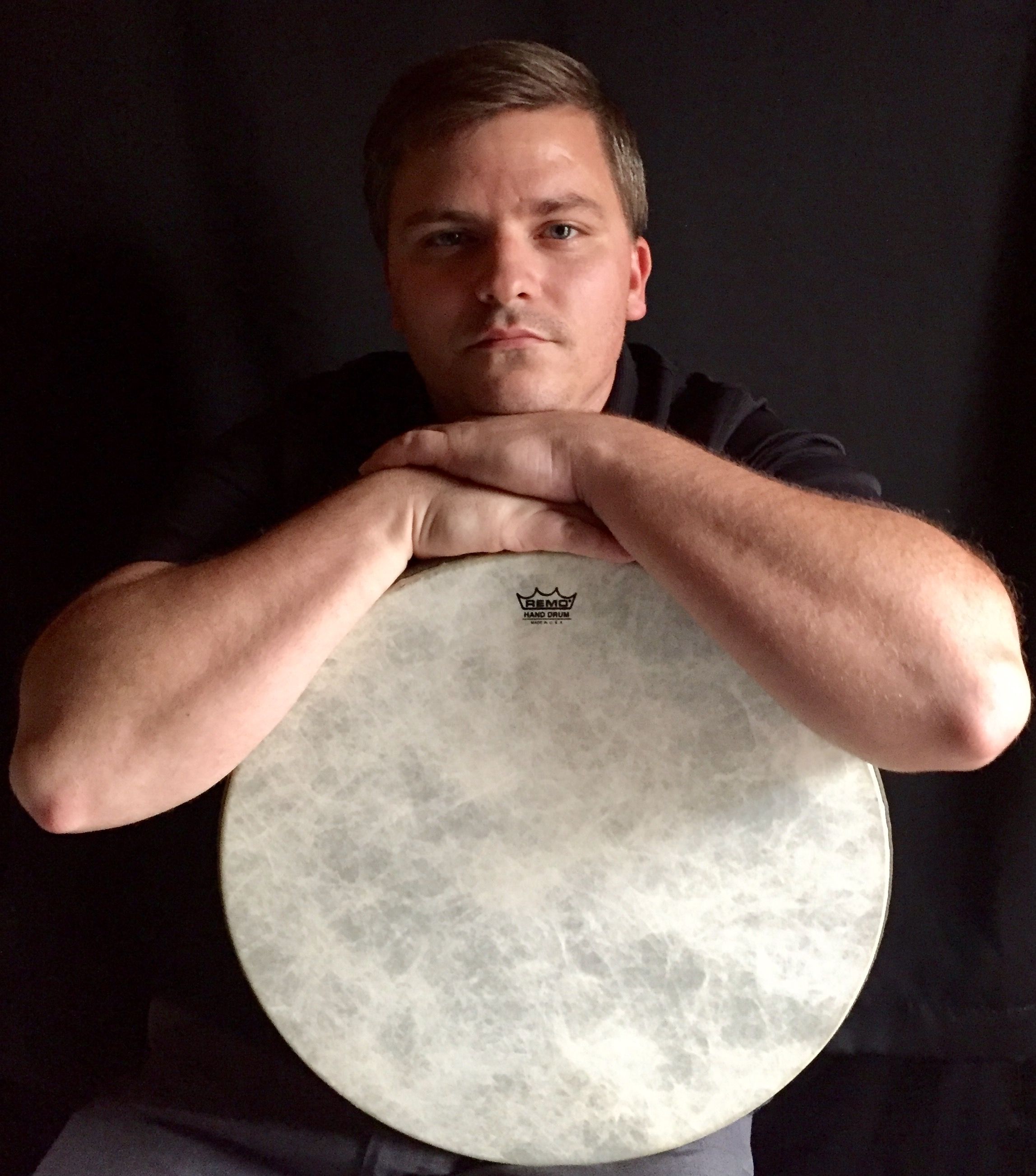 Founder of the Rhythm and Grooves Academy rests his crossed arms on top of a hand drum, looking directly at the camera against a dark background.