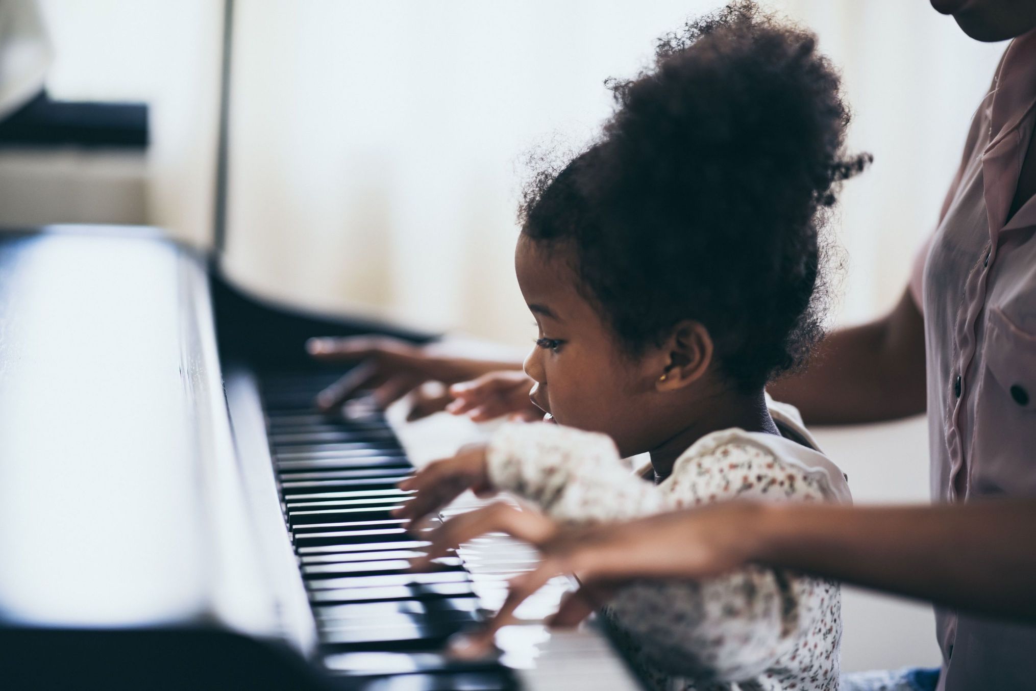 A young girl with curly hair plays piano, closely watching her hands on the keys, with an adult's hand also visible guiding alongside hers.