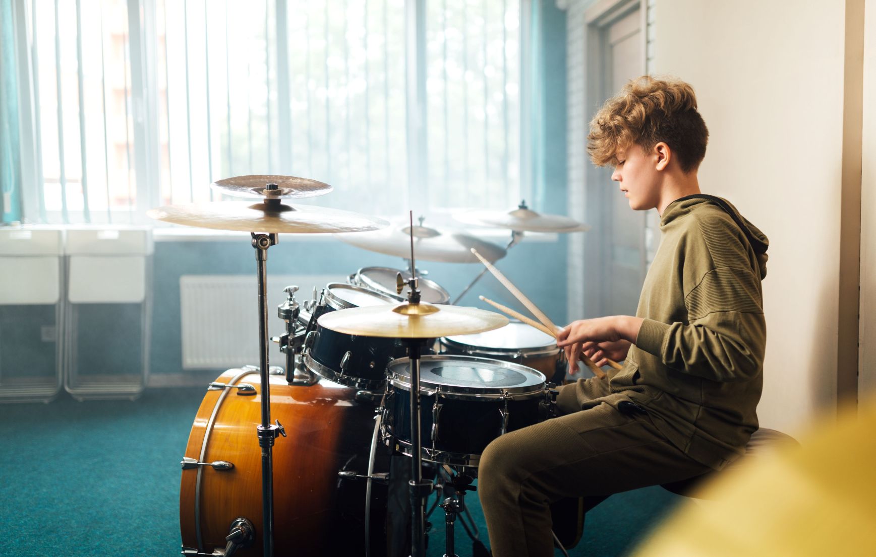 A teenage boy with curly hair, wearing an olive hoodie, sits at a drum kit holding drumsticks in a bright room with large windows.