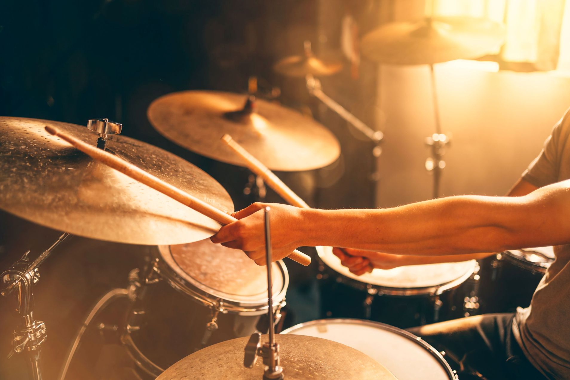 Close-up of a drummer's arm and hands holding drumsticks over cymbals and drums, backlit with warm golden light.