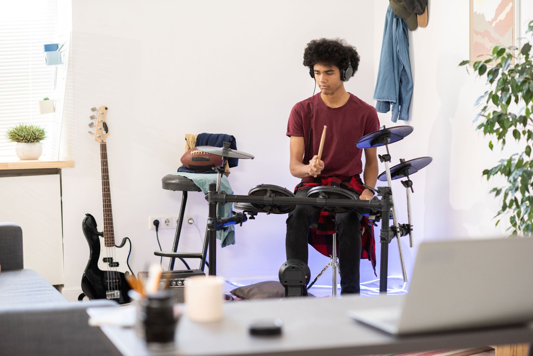 A teenage boy wearing headphones and a maroon shirt plays an electronic drum kit in a bedroom, with a bass guitar leaning against the wall nearby.